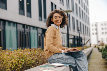 Wall Mural - Smart young woman student in eyeglasses is using pc laptop sitting outdoors on building background