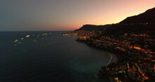 Aerial Backward Shot Of Illuminated Ships In Sea By Residential City By Mountains At Night - Monte Carlo, Monaco