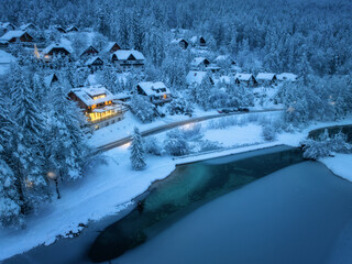 Wall Mural - Aerial view of fairy village in snow, road, forest, Jasna lake, houses, street lights at winter night. Top view of alpine mountains, illumination, snowy pine trees at twilight. Kranjska Gora, Slovenia