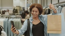 Medium Shot Of Attractive Young African American Woman Video Chatting On Smartphone With Friend While Doing Clothes Shopping In Designer Boutique, Showing Paper Shopping Bags In Her Hands At Camera