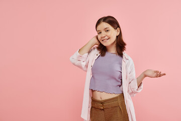 Wall Mural - joyful teenage girl in stylish casual attire pointing with hand and looking at camera on pink