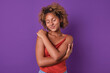 Young happy teenage African American woman dressed in casual clothes hugs herself to cheer up and get rid of feeling of loneliness due to absence of parents nearby stands on purple background