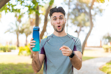 Wall Mural - Young handsome sport man with a bottle of water at outdoors surprised and pointing front