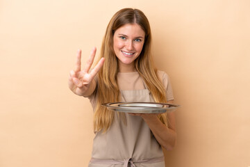 Wall Mural - Young blonde waitress woman with tray isolated on beige background happy and counting three with fingers