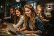 © apratim - Young smiling woman working on laptop at a busy cafe with friends in the background.