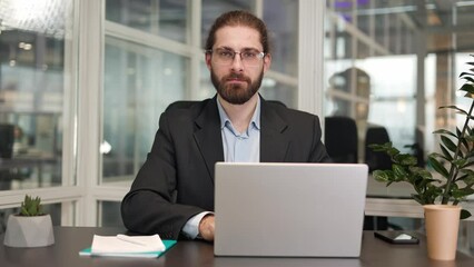 Portrait of stylish creative designer typing personal business plan on modern laptop in office interior. Handsome caucasian worker dressed in formal attire looking directly and smiling indoors.