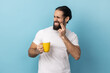 Portrait of man with beard wearing white T-shirt standing and touching his cheek because feeling pain on tooth, after drinking cold water. Indoor studio shot isolated on blue background.
