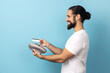 Side view of joyful man with beard wearing white T-shirt using pos payment terminal, going to make payment with card, cashless purchases. Indoor studio shot isolated on blue background.