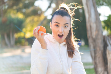 Wall Mural - Young pretty brunette woman holding an orange at outdoors with surprise and shocked facial expression