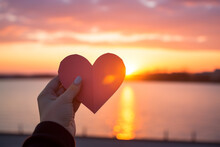 Hands Holding A Paper-cut Heart Shape Against A Sunset Background