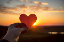Hands Holding A Paper-cut Heart Shape Against A Sunset Background