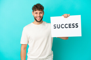 Wall Mural - Young handsome caucasian man isolated on blue background holding a placard with text SUCCESS with happy expression