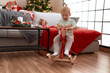 Adorable toddler playing on reindeer rocking by christmas tree at home