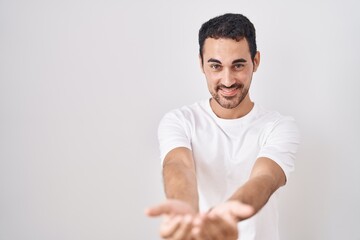 Wall Mural - Handsome hispanic man standing over white background smiling with hands palms together receiving or giving gesture. hold and protection