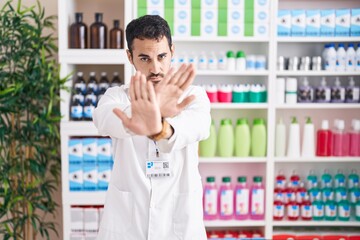 Sticker - Handsome hispanic man working at pharmacy drugstore rejection expression crossing arms and palms doing negative sign, angry face