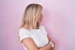 Young blonde woman standing over pink background looking to the side with arms crossed convinced and confident