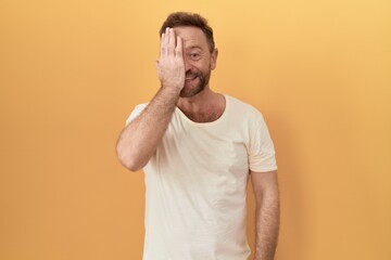 Wall Mural - Middle age man with beard standing over yellow background covering one eye with hand, confident smile on face and surprise emotion.