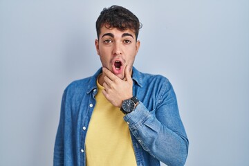 Sticker - Young hispanic man standing over blue background looking fascinated with disbelief, surprise and amazed expression with hands on chin