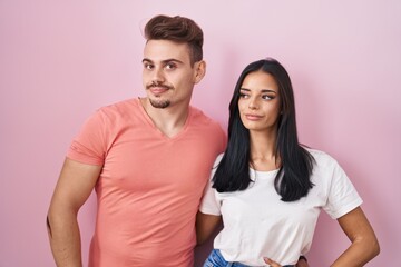 Wall Mural - Young hispanic couple standing over pink background smiling looking to the side and staring away thinking.