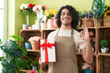 Sticker - Hispanic man with curly hair working at florist shop holding gift doing ok sign with fingers, smiling friendly gesturing excellent symbol