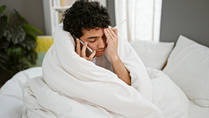 Poster - Young latin man covering with blanket holding thermometer talking on smartphone at bedroom