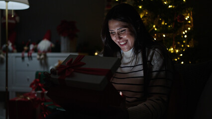 Young beautiful hispanic woman unpacking christmas gift with surprised face at home