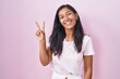 Young hispanic woman standing over pink background smiling with happy face winking at the camera doing victory sign with fingers. number two.