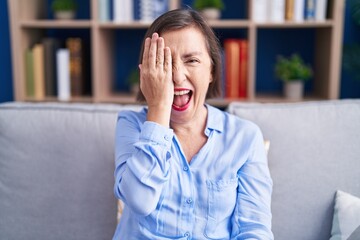 Poster - Middle age hispanic woman sitting on the sofa at home covering one eye with hand, confident smile on face and surprise emotion.