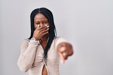 Sticker - African woman with braids standing over white background laughing at you, pointing finger to the camera with hand over mouth, shame expression