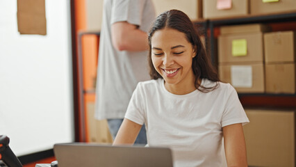 Wall Mural - Confident team of two workers, a man and woman, laughing together while working on a laptop in the vibrant office, packaging ecommerce orders for shipping.