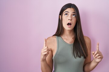 Poster - Hispanic woman standing over pink background amazed and surprised looking up and pointing with fingers and raised arms.