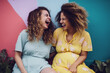 Two pregnant women in summer pastel color dress sitting outside and laughing. Colorful wall background.