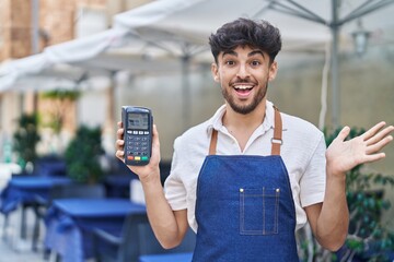 Wall Mural - Arab man with beard wearing waiter apron at restaurant terrace holding dataphone celebrating achievement with happy smile and winner expression with raised hand