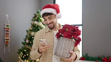 Wall Mural - Young arab man using smartphone holding plant standing by christmas tree at home