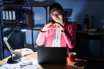 Wall Mural - Chinese young woman working at the office at night smiling in love doing heart symbol shape with hands. romantic concept.