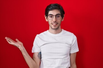 Canvas Print - Young hispanic man standing over red background smiling cheerful presenting and pointing with palm of hand looking at the camera.