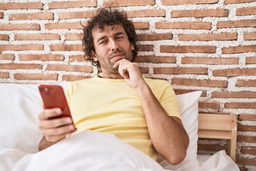 Canvas Print - Hispanic young man using smartphone on the bed serious face thinking about question with hand on chin, thoughtful about confusing idea