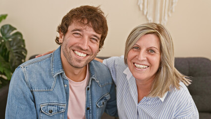 Wall Mural - Heartwarming mother and son sitting together on the living room sofa, joyfully hugging and smiling in their cozy home, radiating happiness and confidence in their casual lifestyle.