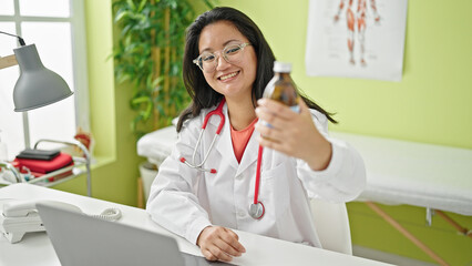 Wall Mural - Young chinese woman doctor using laptop holding medication bottle at the clinic