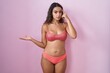 Young hispanic woman wearing lingerie over pink background confused and annoyed with open palm showing copy space and pointing finger to forehead. think about it.