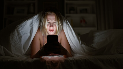 Wall Mural - Young blonde woman using smartphone covering with blanket surprised at bedroom