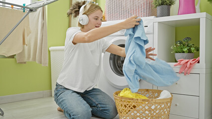 Wall Mural - Young blonde woman listening to music holding clothes of basket at laundry room