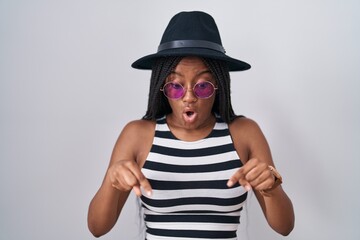 Poster - Young african american with braids wearing hat and sunglasses pointing down with fingers showing advertisement, surprised face and open mouth