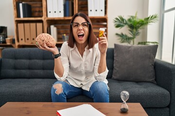 Wall Mural - Young hispanic woman working on depression holding pills angry and mad screaming frustrated and furious, shouting with anger looking up.