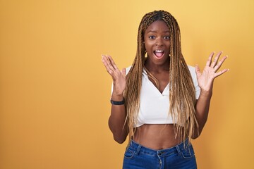 Wall Mural - African american woman with braided hair standing over yellow background celebrating crazy and amazed for success with arms raised and open eyes screaming excited. winner concept