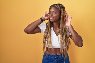 Wall Mural - African american woman with braided hair standing over yellow background trying to hear both hands on ear gesture, curious for gossip. hearing problem, deaf