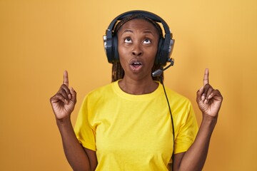 Wall Mural - African american woman listening to music using headphones amazed and surprised looking up and pointing with fingers and raised arms.