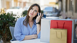 Fototapeta Pomosty - Young beautiful hispanic woman listening to voice message by smartphone sitting on table with shopping bags at coffee shop terrace