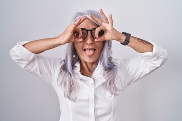 Poster - Middle age woman with tattoos wearing glasses standing over white background doing ok gesture like binoculars sticking tongue out, eyes looking through fingers. crazy expression.