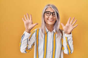 Poster - Middle age woman with grey hair standing over yellow background wearing glasses showing and pointing up with fingers number ten while smiling confident and happy.